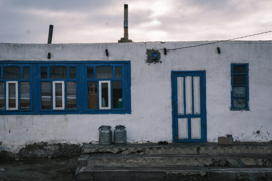 View of a simple white house with blue window frames and doors, featuring metal milk cans on the concrete steps under a cloudy sky in Chah Ab, Gorno-Badakhshan Auto, Tajikistan.
