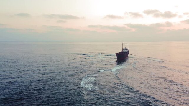 Dilapidated cargo ship adrift on calm ocean waters under a hazy sky, leaving a wake. The vessel leaves a visible wake behind it.