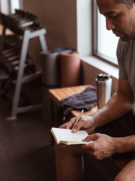 A man in his early thirties sits on a bench inside a gym. He writes in a notebook while looking focused. There are weights and exercise equipment in the background