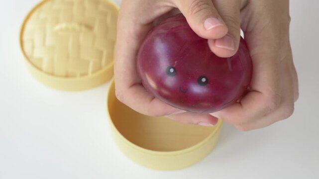 Hand presses glossy red squishy toy closeup revealing soft rubbery deforming texture inside box with steady hand. Sensory antistress gift for collectors highlights charming smiling character face