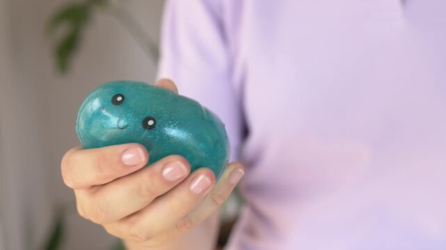 Hand pokes glossy teal squishy character toy closeup revealing soft deforming rubbery texture indoors calmly. Playful tactile press motion highlights collectible sensory toy for stress relief 