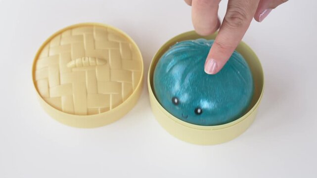 Finger gently presses blue squishy dumpling inside yellow container, demonstrating soft body and glossy texture. Close footage focuses on tactile stimulation, sensory play and fidget activity.
