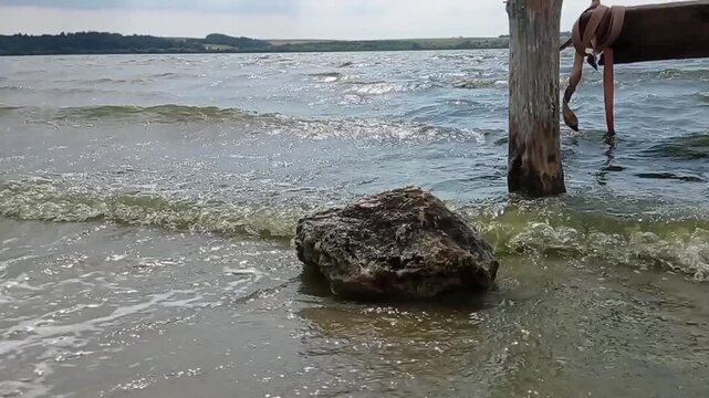 The shore of a pond, with waves rolling onto a sandy beach featuring a large stone.