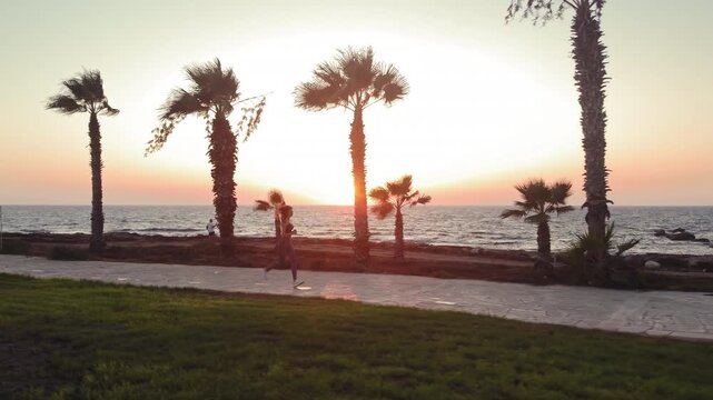 Woman jogs along paved path beside ocean with palm trees during golden hour sunset. The runner moves along the seaside path at sunset.