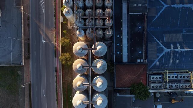 Overhead perspective of large cylindrical industrial storage tanks arranged in rows beside a road and warehouse rooftop, illuminated by warm golden morning light