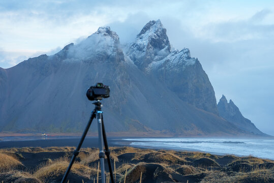 View of a camera on a tripod facing the dramatic Vestrahorn mountain with snow-capped peaks, black sand dunes, and the ocean in Iceland.