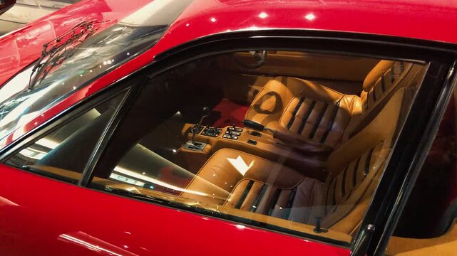 Interior view through the window of a classic red vintage sports car showcasing elegant tan leather Daytona bucket seats, manual gated gear shifter, and vintage dashboard design details.

