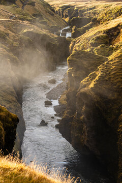 View of a river flowing through a deep, mossy canyon with mist rising from the water under golden hour light in Iceland.