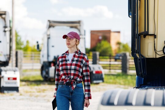 Woman trucker in plaid shirt and cap stands by semi-trucks at depot