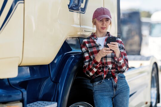 Young female truck driver using smartphone while leaning on semi-truck