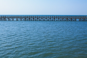 Fototapeta premium Long wooden bridge stretches across calm turquoise water. Traditional Indonesian design, serene seascape, clear blue sky create peaceful tropical atmosphere, ideal for travel and tourism promotions.