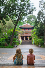 Two children sit on steps facing a traditional Vietnamese temple surrounded by lush tropical greenery in Saigon. Peaceful moment in cultural setting, perfect for family travel, and lifestyle themes. © Tropical studio