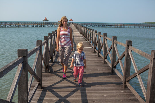 Family with children walks along a long wooden pier stretching into the blue sea on sunny day. Clear blue sky, calm ocean create peaceful, tropical atmosphere. Travel with kids, summer, beach vacation