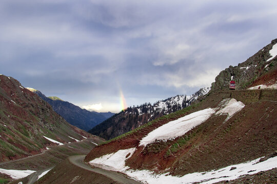 View of the winding road at Lowari Pass with snow-covered slopes, red rocky terrain, and a rainbow appearing in the cloudy sky in Khyber Pakhtunkhwa, Pakistan.