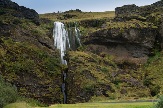 View of a cascading waterfall flowing through a narrow rocky gorge covered in green moss and grass in Eyjafjallajokull, Rangarthing eystra, Iceland.