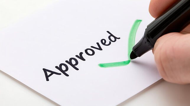 Close up view of human hand hold black marker and draw green check mark near word Approved written on paper sheet on clean white desk