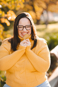 Smiling beautiful woman 30-35 year old wearing yellow knitted sweater and eye glasses holding leaf over autumn leaves close up. Fall season.