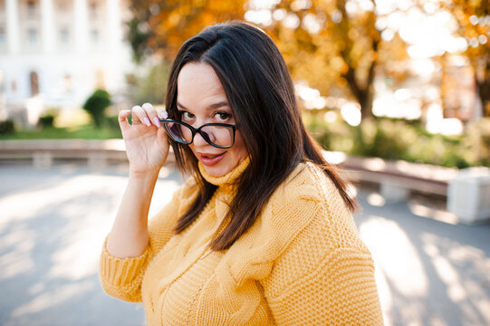 Smiling beautiful woman 30-35 year old wearing yellow knitted sweater and eye glasses over autumn leaves close up. Fall season. Looking through at camera.