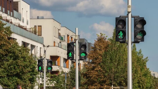 Timelapse of pedestrian and bicycle traffic lights changing from red to green at an urban intersection, with modern residential buildings and contemporary city architecture in the background