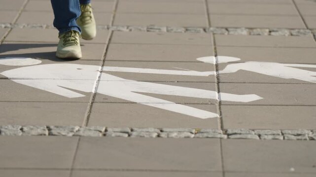 Close up of pedestrian walking toward the camera over painted pedestrian path marking, feet in sneakers approaching along designated urban walkway on sunny summer day