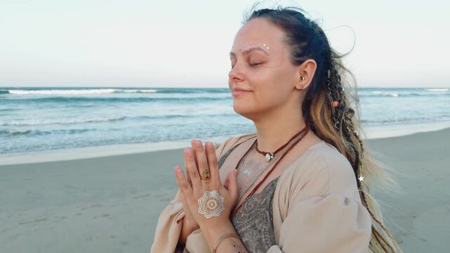 Woman standing with hands in namaste gesture and eyes closed on a sandy beach, focusing on spiritual meditation and calm mindfulness with the ocean waves in the background