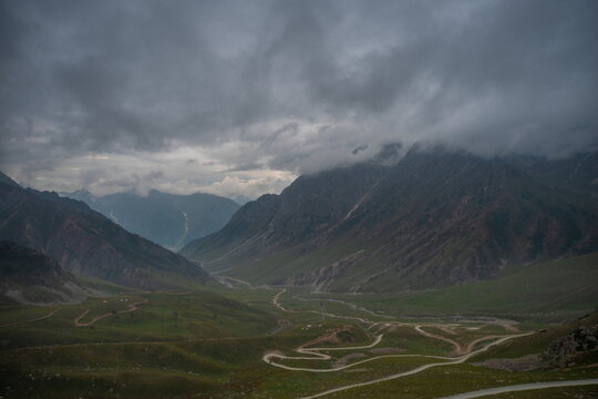 View of the winding road through the lush green valley of Burzil Pass under a dramatic cloudy sky in Astore, Gilgit Baltistan, Pakistan.