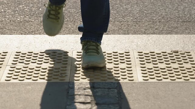 Close up front view pedestrian feet stepping on tactile warning paving at urban crosswalk, person in jeans and sneakers crossing street on sunny day, road markings and accessible city infrastructure