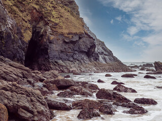 Dark, rugged cave entrance framed by weathered rock layers. Rough, uneven stones scattered near opening, rough ocean on the right. Perfect for backgrounds, adventure themes © mark_gusev