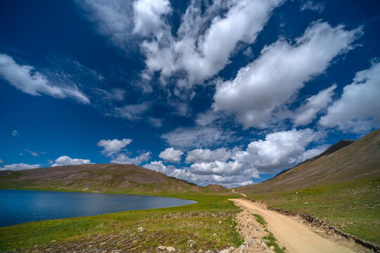 View of Sheosar Lake surrounded by rolling green hills and a dirt road under a bright blue sky with fluffy clouds in Deosai National Park, Gilgit Baltistan, Pakistan.