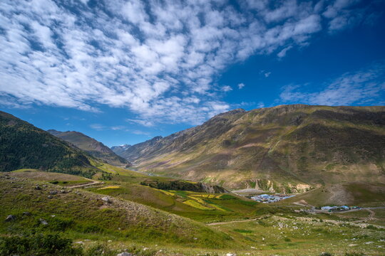 View of Deosai National Park with rolling green hills, rugged mountain slopes, a small settlement in the valley, and a bright blue sky in Astore, Gilgit Baltistan, Pakistan.