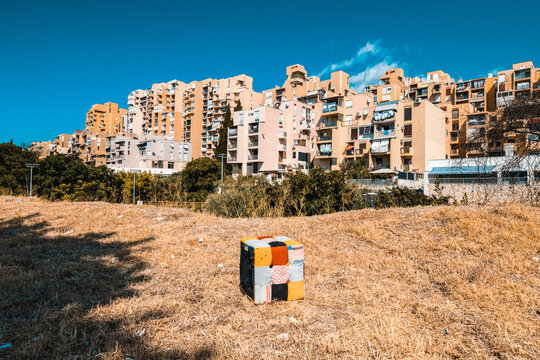 View of a colorful patchwork cube sitting in a dry grassy field in front of large residential apartment buildings in Split, Split-Dalmatia County, Croatia.