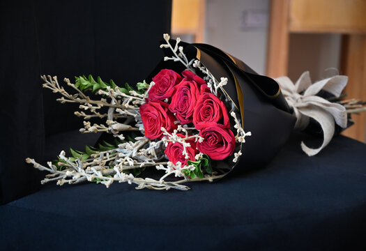 elegant red roses bouquet in black wrapping paper with white ribbon, romantic flower gift resting on dark chair, floral design close up