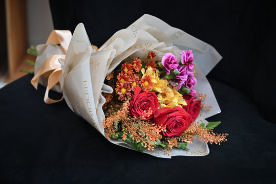 vibrant flower bouquet wrapped in white tissue paper, floral arrangement with red roses, purple carnations and yellow alstroemeria resting on dark surface, gift bouquet close up