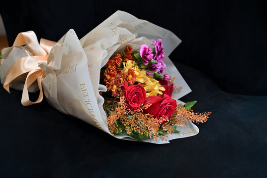 vibrant flower bouquet wrapped in white tissue paper, floral arrangement with red roses, purple carnations and yellow alstroemeria resting on dark surface, gift bouquet close up