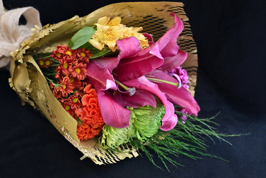 vibrant flower bouquet wrapped in craft paper resting on dark surface, colorful floral arrangement with pink lilies, orange celosia and green chrysanthemum, gift bouquet close up