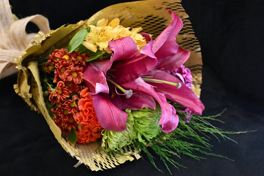 vibrant flower bouquet wrapped in craft paper resting on dark surface, colorful floral arrangement with pink lilies, orange celosia and green chrysanthemum, gift bouquet close up