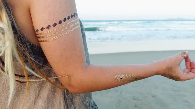 Woman performing a mudra gesture with her hand, adorned with gold temporary tattoos, engaging in a peaceful meditation practice on a sandy beach by the ocean
