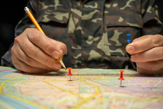 A person places red and blue pins on a map while holding a pencil. The setting is dim, and the individual wears a camouflaged shirt. The map shows various streets and areas