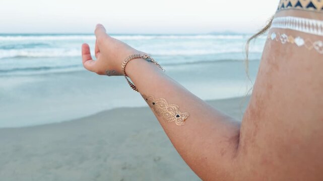 Woman practicing yoga and meditation pose with mudra hand gesture, wearing temporary metallic tattoos on arm and shoulder, finding peace and mindfulness on a calm beach