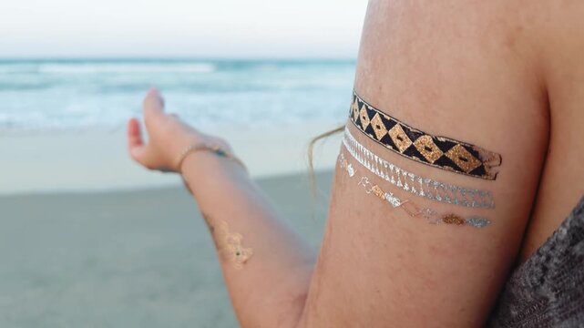 Woman holding an open palm gesture with ornate metallic temporary tattoos on her arm, symbolizing a serene moment of meditation and connection with nature at the tranquil beach