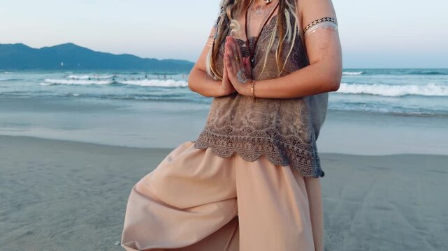 Woman standing on a sandy beach at sunset, performing a yoga or meditation pose with hands in namaste mudra, embodying mindfulness, spiritual balance, and a serene lifestyle by the ocean