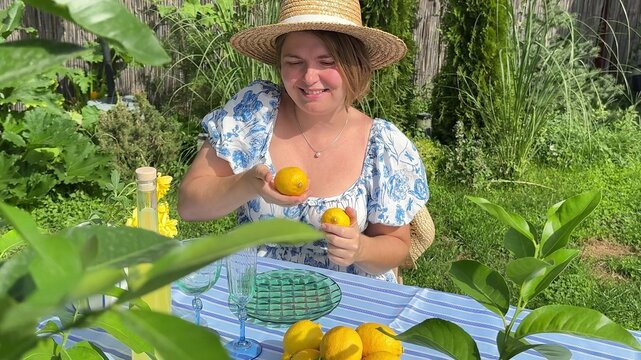 Happy Caucasian woman in straw hat and floral dress, holding fresh lemons at a garden table, sunny summer day, rustic outdoor setting