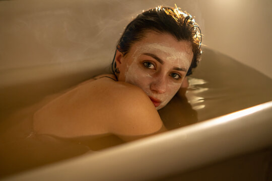 Young woman in bathtub with cosmetic clay face mask, warm bath steam, looking at camera