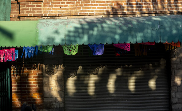 View of colorful papel picado decorations hanging from a green awning above a closed storefront with a brick wall in Oaxaca, Oaxaca, Mexico.