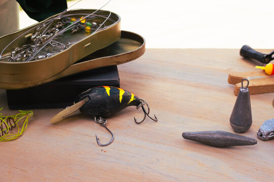 Fishing tackle spread out on a light wooden surface, with a lure, several weights and a metal box full of hooks and swivels, ready for a day of fishing outdoors.