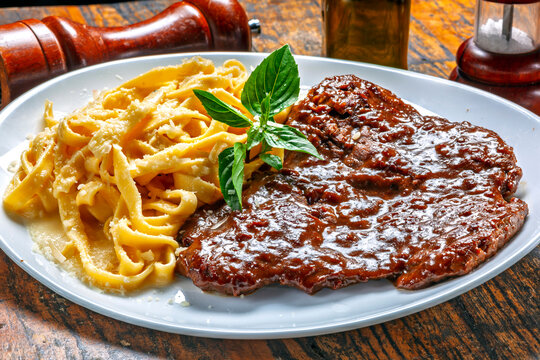 Delicious butterfly steak in rich sauce with creamy fettuccine alfredo pasta and fresh basil on a white plate over a rustic wooden table.