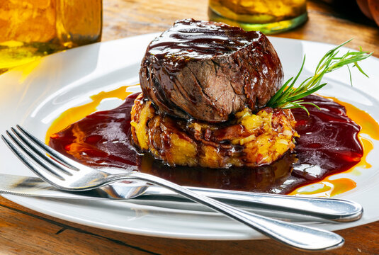 Close-up photo of a grilled beef filet mignon on a sweet potato hash bed, with red wine and orange sauces, garnished with fresh rosemary. White plate, silverware
