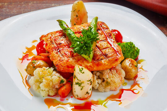Gourmet grilled salmon fillet with roasted vegetables, asparagus, broccoli, and savory sauce on a white plate.