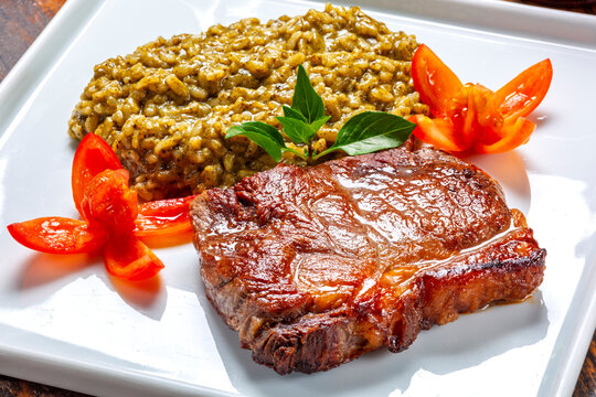 Juicy grilled steak paired with creamy pesto risotto, garnished with fresh basil and tomato flowers on a white plate.