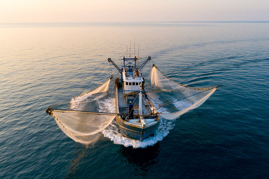 Aerial view of a commercial fishing boat trawler with large open nets moving in the blue sea water at sunset. Industrial marine fishery and seafood catch concept